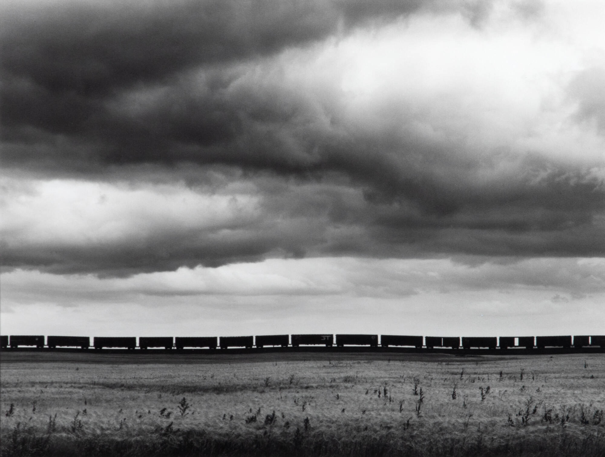 Black and white photograph of a silhouette of train cars traveling across large open field, stormy sky.